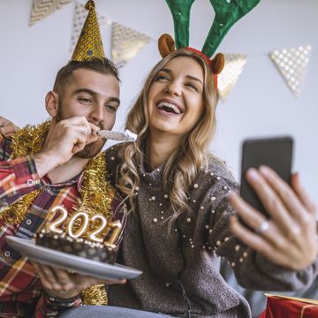 couple celebrating christmas at home, holding chocolate cake with lit candles shaped as numbers 2021 and having a video call with friends