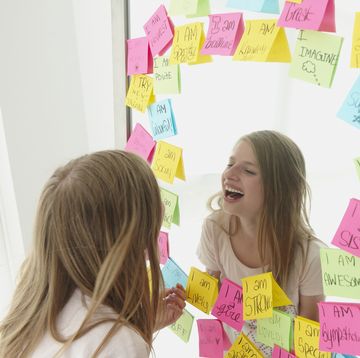 caucasian girl laughing in mirror with positive affirmation adhesive notes around the mirror