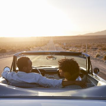 rear view of couple on road trip driving classic convertible car towards sunset